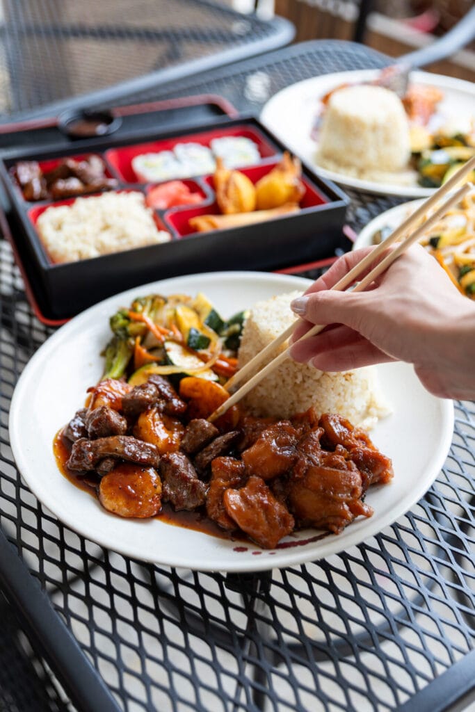 Hibachi dish surrounded by other plates of food, someone picking up meat with chopsticks