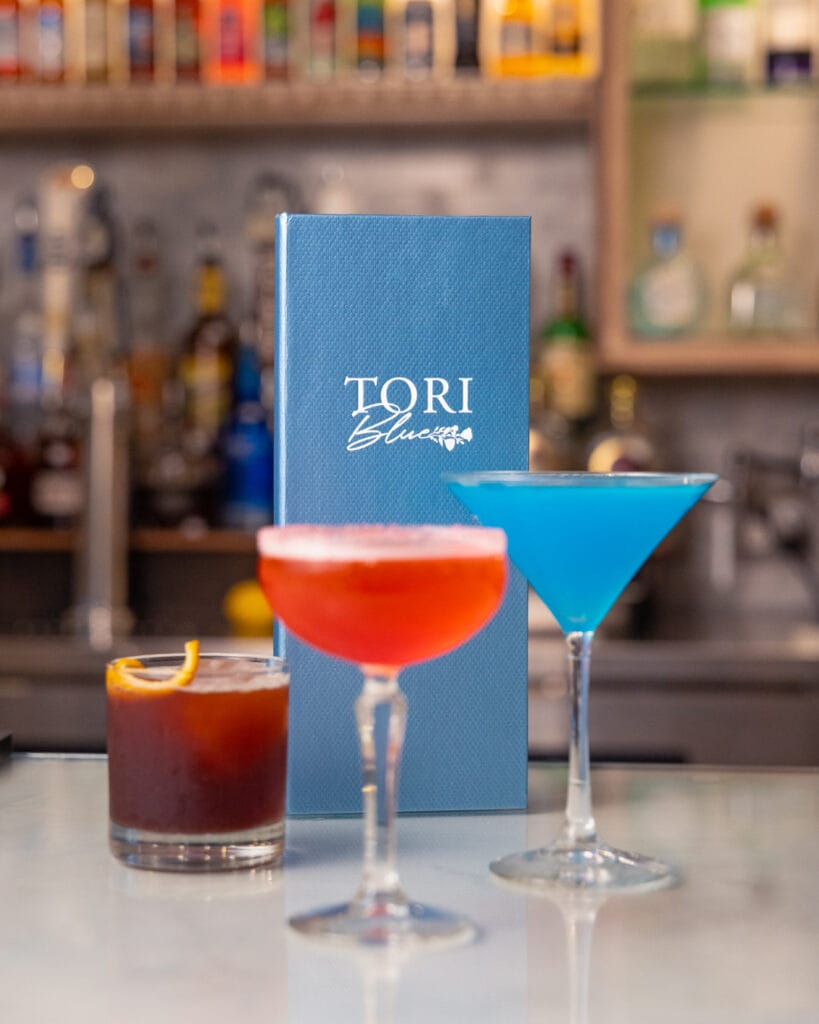 Colorful cocktail drinks sitting on top of bar in front of blue Tori Blue menu